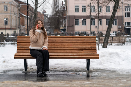Young woman sits on park bench in city and talks on phone . Girl in beige sheepskin coat rests outside in winterの写真素材