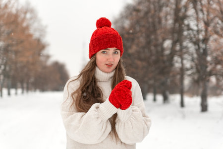 Portrait of beautiful young woman in red knitted hat and mittens and woolen sweater on alley in winter parkの写真素材