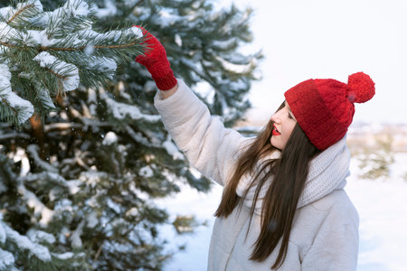 Portrait of brunette girl in red knitted hat near the snow-covered fir trees. Young woman walking in forest.の写真素材