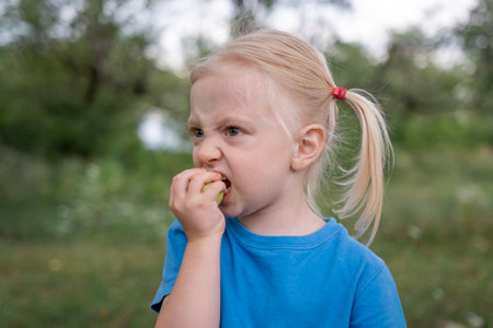 Fair-haired little girl in blue t-shirt eats green apple outside. Child walks in the park or garden. Close-up portrait of little girlの写真素材