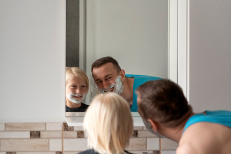 Young dad and little boy with shaving foam on face look in bathroom mirror. Dad and son shave and have fun.の写真素材