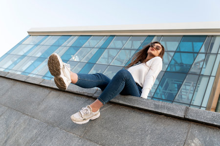 Attractive stylish woman resting on city street.Young brunette girl sits on parapet with her legs hanging down.Urban.の写真素材