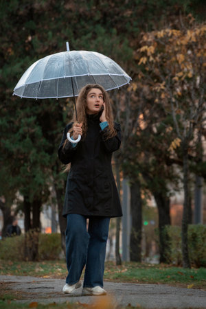 Woman with transparent umbrella walking in park in fall and talking to phone. Young beautiful woman using smartphone under umbrellaの写真素材