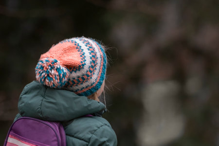 Back view child in knitted wool hat and bologna jacket with small backpack outdoors. cold autumn day
