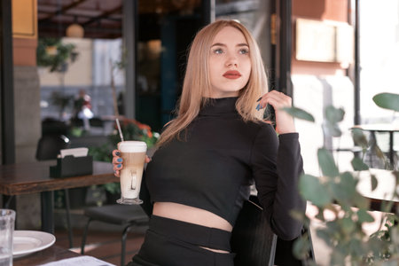 Brooding attractive young blonde woman sits at table in coffee shop with cup of cappuccino coffee latteの写真素材