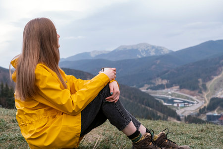 Female traveler looks at the mountains. Tourist girl sits on hillside with cup in autumnの写真素材
