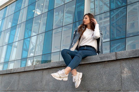 Stylish young woman resting on summer city street. Beautiful student girl in jeans sitting near modern building.の写真素材