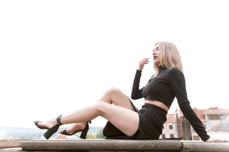 Beautiful slender blonde girl in black clothes wears heels sitting on observation deck and showing off her beautiful legsの写真素材