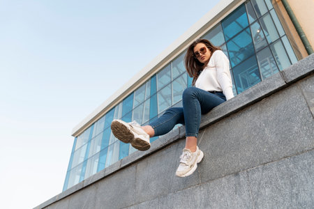 Young brunette girl sits on the parapet with her legs hanging down. Urban. Attractive stylish woman resting on city streetの写真素材
