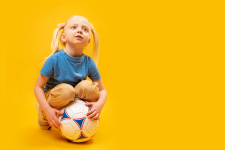 Studio photo of small caucasian white-haired girl with two tails holds soccer ball in isolation on yellow background. copy spaceの写真素材