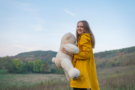Beautiful long-haired girl laughs and plays with teddy bear on autumn day. Young woman in yellow park jacket walks in nature.の写真素材