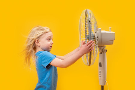 Little blonde girl with windblown hair near fan on yellow background. Fair-haired child near blowerの写真素材