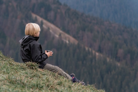 Young fair-haired boy sits on hillside with cup of tea. Portrait of child in mountains. Travel hiking outdoorの写真素材