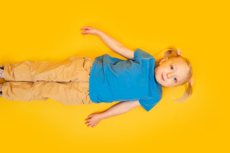 Portrait of preschool girl on bright yellow background. Little girl in blue T-shirt lies on yellow floor. top viewの写真素材