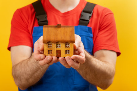 Small toy wooden house in mens hands. Man in worker uniform holds model at home Close up.の写真素材