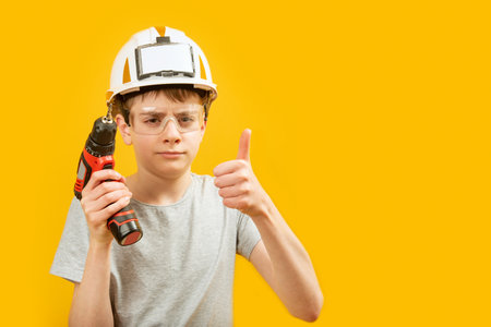 Teenage boy wears protective helmet and glasses holding drill and showing thumbs up isolated on yellow background. child as builder. copyspace.の写真素材