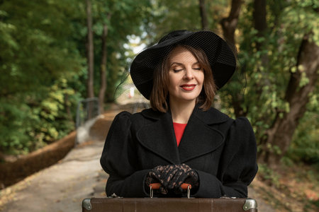 Portrait of happy beautiful woman with closed eyes in black hat, classic jacket and gloves, hands on...