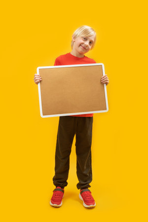 Vertical frame full-length portrait of blond boy with cortical board in his hands isolated on yellow background. Place to advertise. copy spaceの写真素材