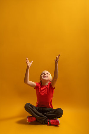 Vertical frame portrait of caucasian blond boy sitting cross-legged and arms up on orange background. copy spaceの写真素材