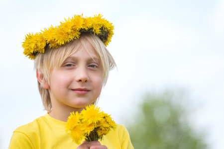 Portrait of boy with white hair wreath of flowers holds bouquet of dandelions looks at the camera and smilesの写真素材
