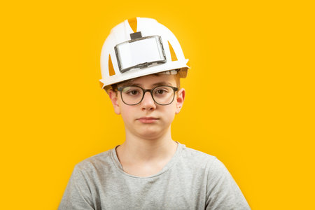 Portrait of teenage boy wearing glasses and protective white helmet isolated on yellow background. Child is like builder or an engineerの写真素材