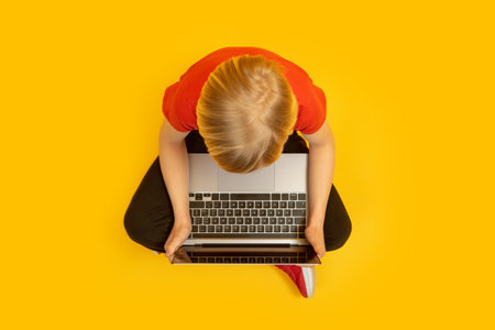 Blond boy sits cross-legged and studies or plays games at laptop isolated on yellow background. top viewの写真素材