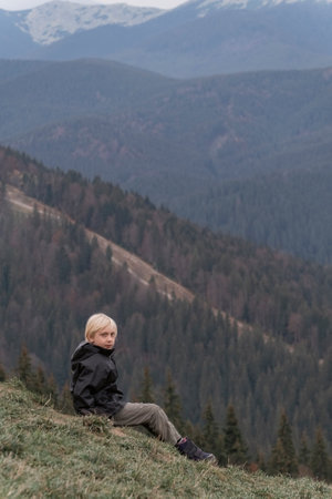 Blond schoolboy sits at hillside and looks at the camera. Child on vacation in mountain. vertical frame.の写真素材