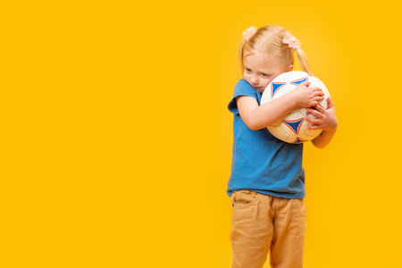 Little blonde girl hugs soccer ball. yellow studio background. copy space mockup. Football for kidsの写真素材
