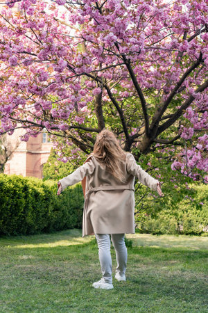 Young woman enjoying warm spring day in park near cherry blossoms. Back view portrait of girl near flowering tree. vertical frameの写真素材