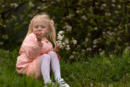 Preschooler girl wearing pink outfit sits on the grass near blooming tree and holds flower. child outdoors in springの写真素材