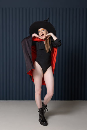 Surprised beautiful young woman in black hat, bodysuit and cloak. Black Friday.Halloween party. Gray background.の写真素材