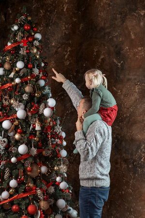 Daughter sits on dads shoulders near beautiful Christmas tree. Portrait of father with little girl on Christmas Eve. New Year moodの写真素材