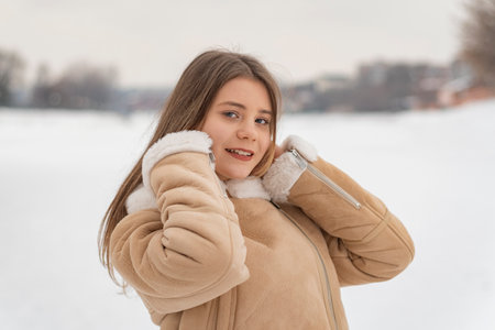 Smiling girl in beige sheepskin coat correcting hair, snowy terrain background. Young woman in winter clothes.の写真素材