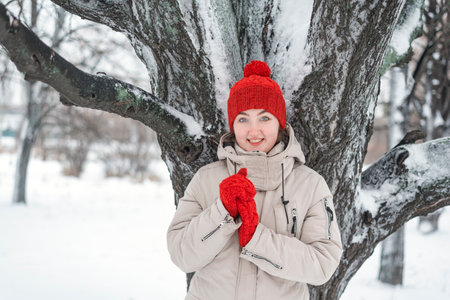 Smiling beautiful young woman under snow-covered tree, looking in the camera. Young woman in red knitted hat and mittens.の写真素材