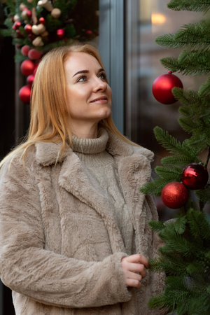 Beautiful red-haired young woman in a fur coat looks up to the Christmas tree, standing on the street. Christmas holidays. Vertical frame.の写真素材