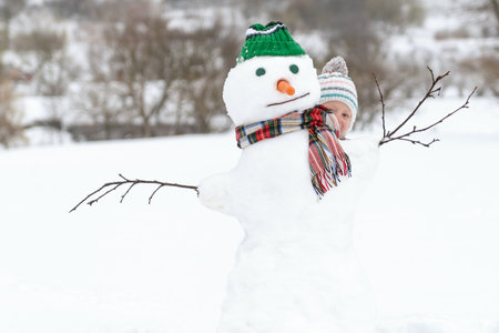 Happy child peeking out behind snowman on winter walk. Winter activities for kids. Kid outside building snow figureの写真素材