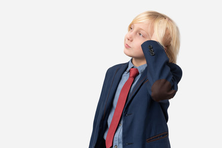 Thoughtful boy with blond hair dressed in blue shirt, red tie and blue jacket standing with hand on chin and looking away isolated over white background.の写真素材