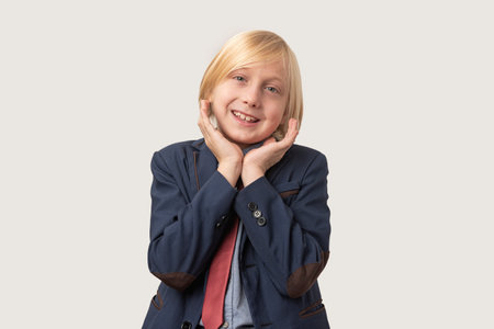 Portrait of little boy with blond hair and blue jacket, keeps hand on cheek, looks directly at camera, isolated over white background. Emotions, facial expressions concept.の写真素材