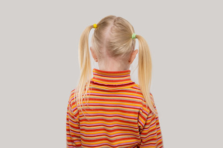 Back view of little blonde girl with ponytail wearing striped shirt isolated on gray background.の写真素材
