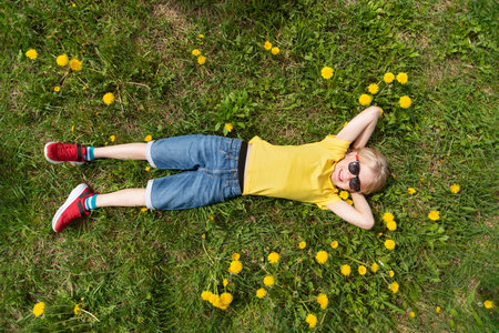 Smiling child lies on grass in dandelions flower field and dreaming. Boy puts his hands behind his head.Top view.の写真素材