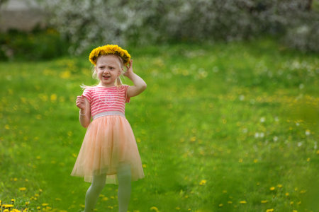 Cute little girl on meadow with flowers. Fair-haired girl wears wreath of dandelions. Summer vacation timeの写真素材