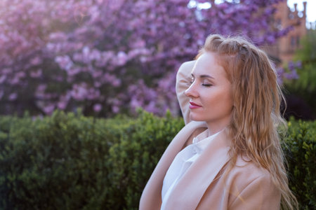 Attractive young woman with closed eyes and red hair in blooming spring park. Cherry blossom tree background. Side viewの写真素材