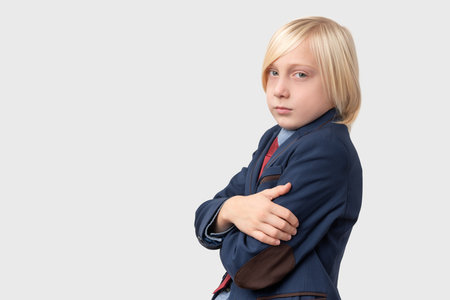 Portrait of serious schoolboy with blond hair, keeps arms crossed and looks directly at camera, isolated over white background.の写真素材