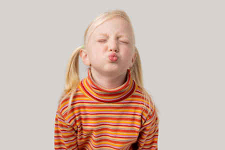Portrait of a cute little girl with blond hair blowing a kiss to the camera with closed eyes, isolated on gray.の写真素材