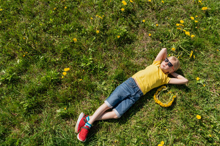 Boy in yellow t-shirt and jeans shorts with hands behind head lying on grass and rest. Countryside. Top viewの写真素材