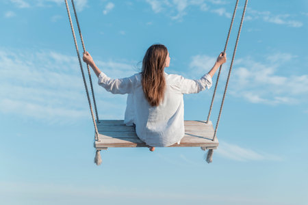 Young woman with long hair on swing high in mid air on blue sky background. Back view. Concept of freedom and lightnessの写真素材