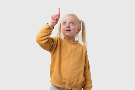 Portrait of a cute little girl with blond hair, wearing a yellow sweater, pointing up, isolated on white backgroundの写真素材
