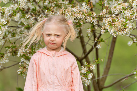 Portrait of frustrated or angry little girl outside near flowering tree with funny grimace on her...