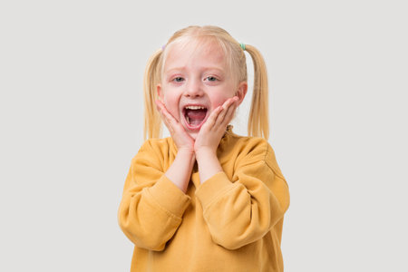 Portrait of a cheerful little girl with blond hair and blue eyes, wearing a yellow sweater, screaming and looking at the cameraの写真素材