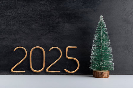 Artificial small Christmas tree on wooden stand and golden number 2025 on black background. New year.の写真素材